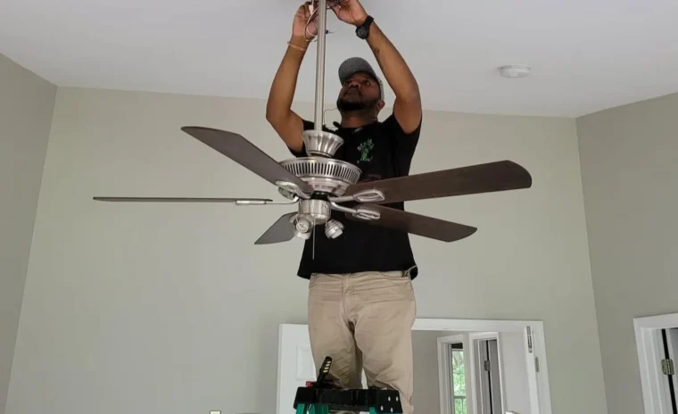 A person wearing a black shirt and beige pants stands on a green ladder, installing or repairing a ceiling fan in a room with light gray walls and white ceiling.